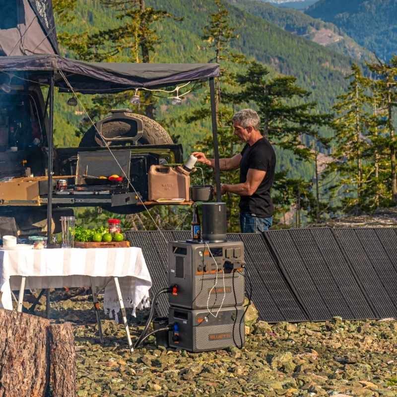 Camper using Bluetti power station for outdoor cooking setup during a mountain camping trip, with solar panels and supplies
