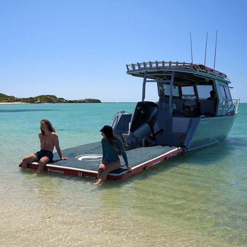 Two people sitting on a Floatables U-Dock inflatable boat dock, attached to a boat in shallow, clear waters, with a scenic island backdrop