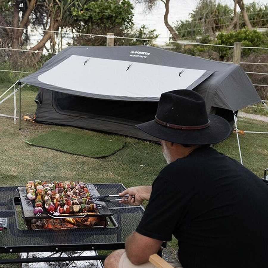 Dometic Pico FTC 2x2 TC-I pitched with the awning up at a coastal campsite while a bloke cooks skewers nearby