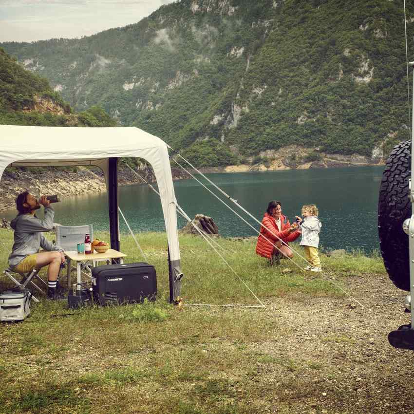 Camp set up under the Dometic Santorini FTK 4x8 TC canopy with a table, chairs and a fridge beside a lake