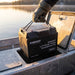 Gloved hand carrying the Renogy Pro 100Ah battery by its rope handle from a small tinnie's under-seat hatch, frost on the hull at dawn
