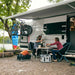 Thule Omnistor 6300 Awning set up at a lakeside campsite, with a couple enjoying coffee under the shade, showcasing the awning's practical use.