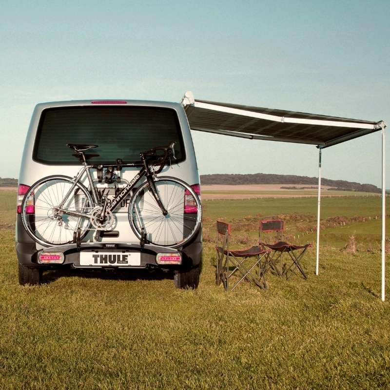 Thule 5102 manual awning extended, providing shelter for a relaxing camping setup next to a vehicle with bikes and chairs.